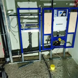 Stelaż podtynkowy Geberit zamontowany w surowym stanie, widoczne rury kanalizacyjne, przewody i poziomica na podłodze z wylewką.