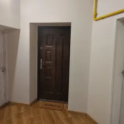 Wnętrze przedpokoju z brązowymi drzwiami wejściowymi, brązowym dywanikiem z napisem 'Dzień dobry!', panelami podłogowymi imitującymi drewno, szarym licznikiem gazu i żółtą rurą gazową na ścianie.