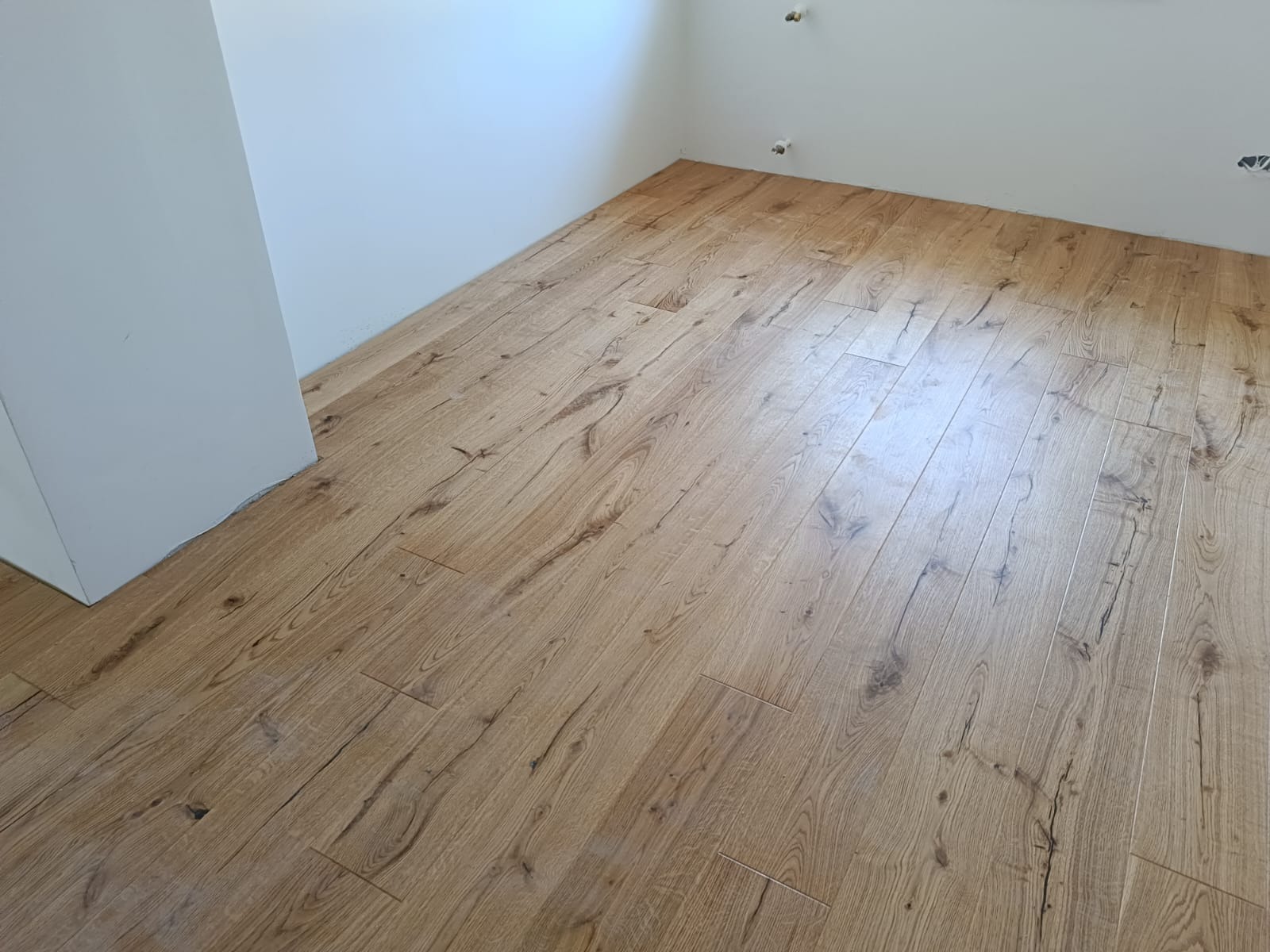 Ułożone panele podłogowe o wzorze drewna w jasnym pomieszczeniu z białymi ścianami, widoczne niedokończone instalacje na ścianie.