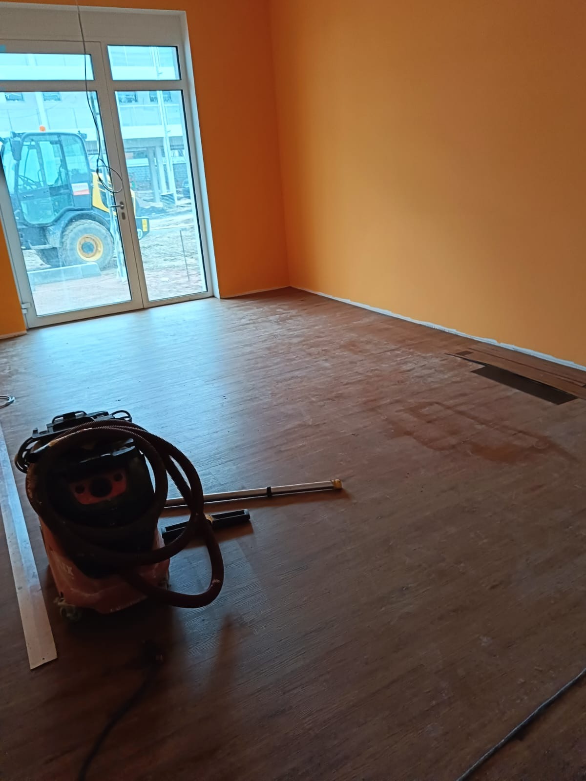 Pomarańczowy pokój z częściowo ułożoną podłogą z paneli, widoczny odkurzacz przemysłowy i fragmenty paneli, za oknem widoczna maszyna budowlana.
