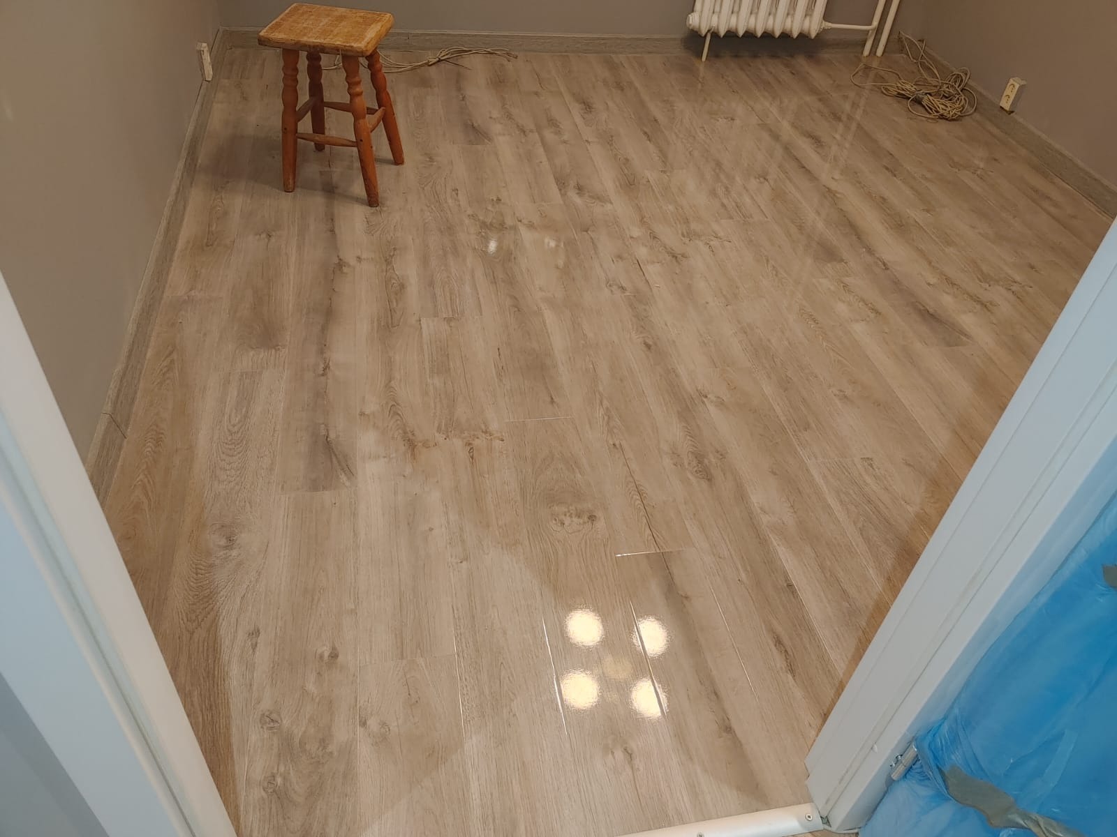 Jasne panele podłogowe imitujące drewno, widok z góry, niewielki stołek drewniany w rogu pokoju, grzejnik pod ścianą