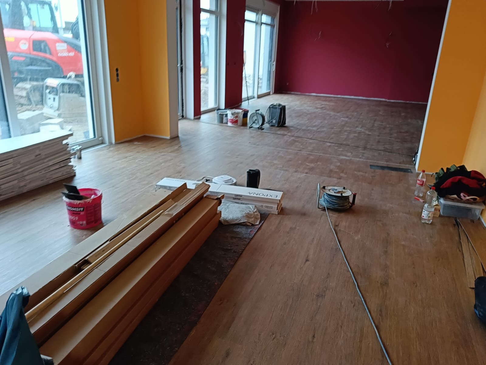 Wnętrze w trakcie remontu: panele podłogowe w kartonach, pomalowane ściany w odcieniach żółci i czerwieni, widoczne narzędzia i akcesoria budowlane, częściowo położona podłoga.