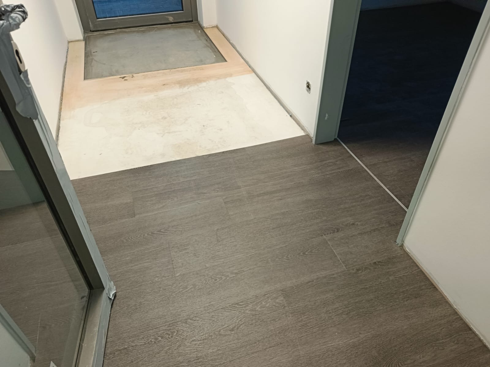 Położona podłoga z paneli w odcieniach szarości, widoczny fragment nieprzygotowanego podłoża i otwór drzwiowy do innego pomieszczenia.