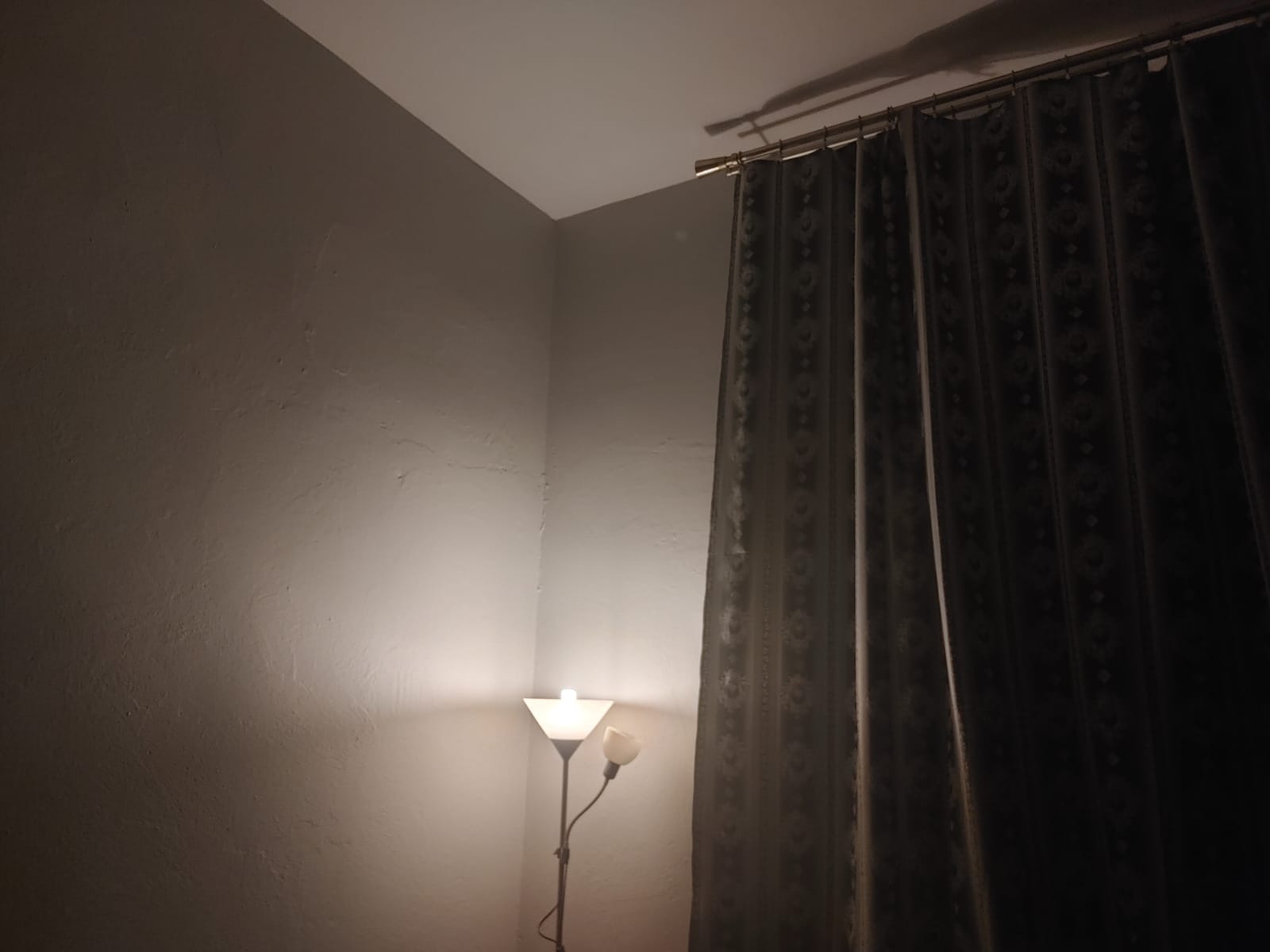 Szary pokój z lampą podłogową oświetlającą ścianę z widoczną fakturą i zasłonami z wzorem kwiatowym przy karniszu.
