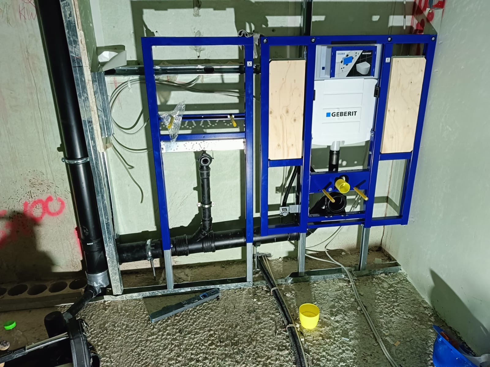 Stelaż podtynkowy Geberit zamontowany w surowym stanie, widoczne rury kanalizacyjne, przewody i poziomica na podłodze z wylewką.