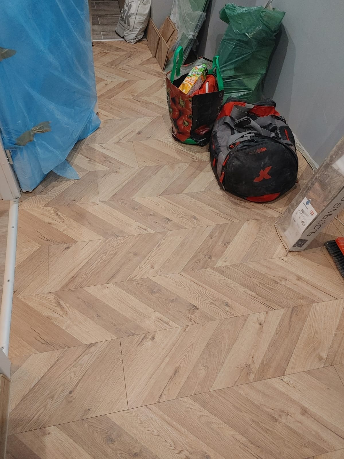 Ułożone panele podłogowe w jodełkę, z torbami i workami w tle, widoczne opakowanie paneli i szczotka do zamiatania.
