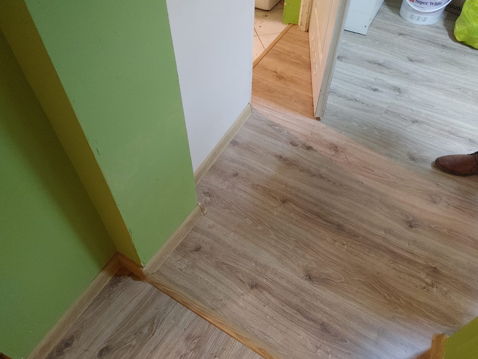 Fragment wykończonego wnętrza z panelami podłogowymi w odcieniach brązu, zielona ściana i biały narożnik, widoczny fragment brązowego buta.
