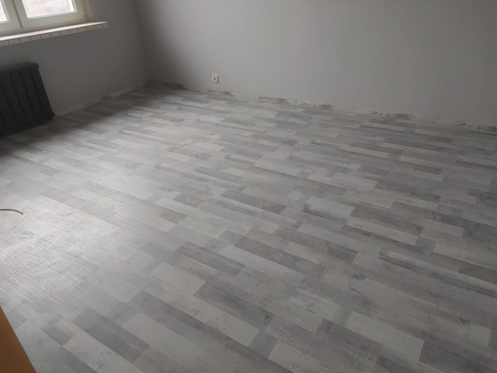 Szare panele podłogowe ułożone w nowym pokoju, widoczny fragment grzejnika i okna.