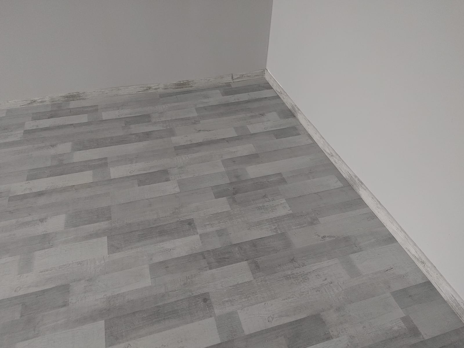Szare panele podłogowe imitujące drewno, ułożone w nowym pomieszczeniu z białymi ścianami i szarą listwą przypodłogową.