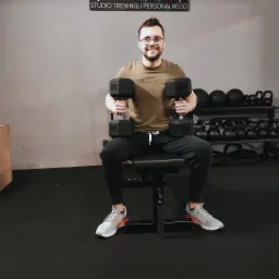 Uśmiechnięty mężczyzna w okularach siedzi na ławce w siłowni, trzymając w obu rękach czarne hantle sześciokątne; w tle regał z kulami i odważnikami.