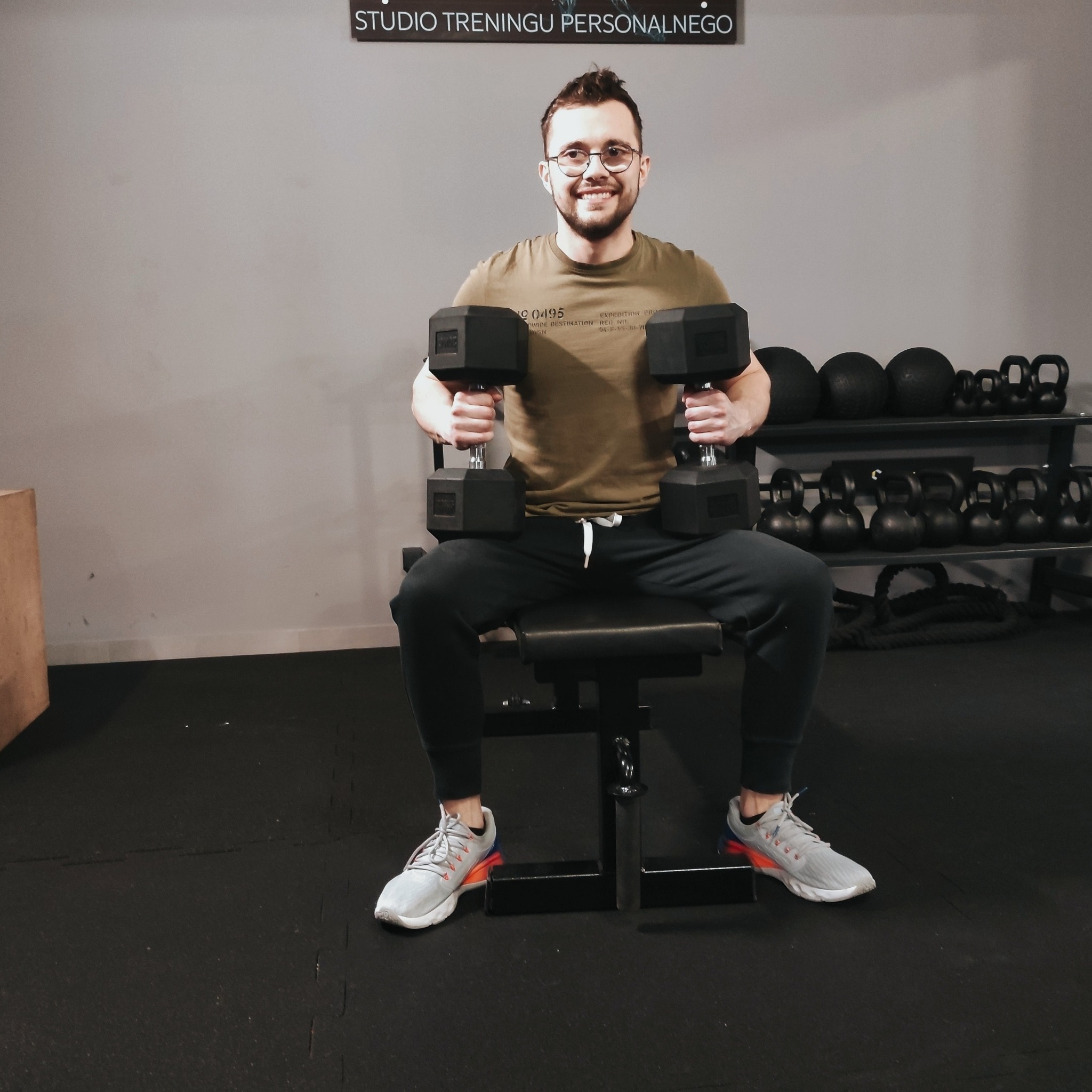 Uśmiechnięty mężczyzna w okularach siedzi na ławce w siłowni, trzymając w obu rękach czarne hantle sześciokątne; w tle regał z kulami i odważnikami.