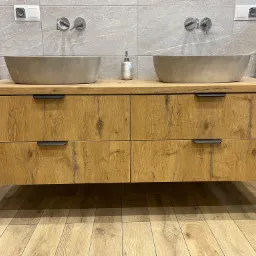 Łazienka z dwoma kamiennymi umywalkami nablatowymi na drewnianej szafce wiszącej z czterema szufladami, srebrny dozownik mydła, szare płytki na ścianie i podłodze imitujące drewno.