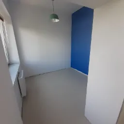 Wnętrze pokoju po malowaniu: ściany w odcieniach bieli i błękitu, szara podłoga, widoczny grzejnik i zielona lampa wisząca.