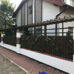 Ocieplony budynek z drewnianą pergolą i ogrodzeniem, ceglane słupki ogrodzeniowe, bluszcz pnący się po ogrodzeniu.