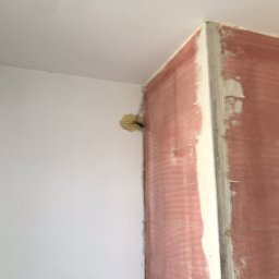 Crado - Ściana narożna w trakcie remontu: fragment ściany pomalowany na różowo, obok biała ściana z widocznym otworem na instalację elektryczną, sufit biały, widoczne ślady szpachlowania.