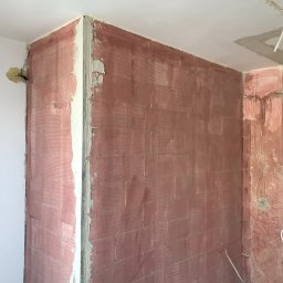 Crado - Narożnik pokoju w trakcie prac przygotowawczych do malowania, ściany pokryte czerwoną warstwą gruntującą, widoczne kable elektryczne i fragment ocieplenia wystający ze ściany.