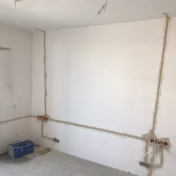 Crado - Przygotowanie ścian do malowania: wyfrezowane bruzdy pod instalację elektryczną, wypełnione gipsem, widoczna poziomica, niebieskie wiadro i wystające przewody elektryczne.