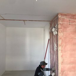 Crado - Praca elektryczna w surowym wnętrzu: instalacja przewodów elektrycznych na ścianie z czerwonej cegły, widoczne kable, poziomica i pracownik w rękawicach.