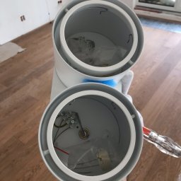 Crado - Widok z góry na białą, podwójną oprawę oświetleniową cylindryczną z widocznymi elementami montażowymi wewnątrz, częściowo rozpakowanymi, trzymaną przez osobę w rękawicy, z widocznym wkrętakiem,...