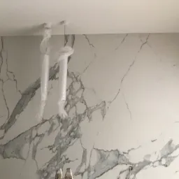 Ściana wyłożona płytami imitującymi biały marmur z szarymi żyłami, z widocznymi dwoma lampami wiszącymi zabezpieczonymi folią ochronną, oraz fragment drabiny u dołu kadru.