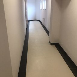 Crado - Korytarz w budynku po remoncie, z jasną podłogą wyłożoną płytkami i ciemnymi listwami przypodłogowymi, pomalowany na jasny kolor z widocznymi dwoma oknami w oddali.