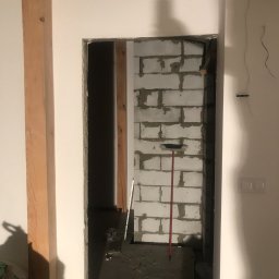 Crado - Wnętrze w trakcie remontu: widok przez otwór drzwiowy na ścianę z bloczków betonowych, z widoczną łatą murarską i miotłą opartą o ścianę, surowy stan wykończenia podłogi i ścian.