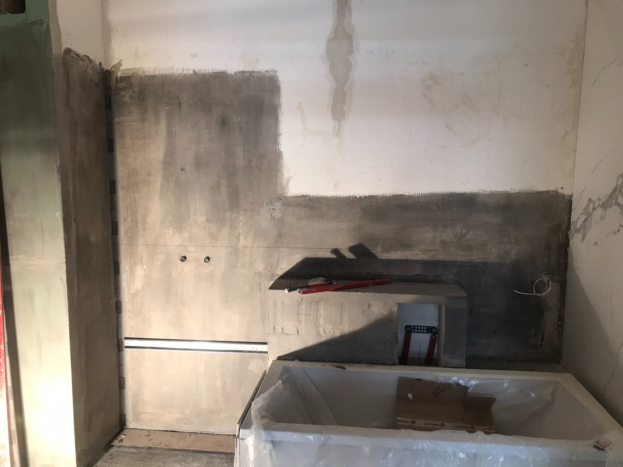 Łazienka w trakcie remontu, widoczne surowe ściany częściowo pokryte szpachlą, przygotowana wanna zabezpieczona folią, poziomica na półce z bloczków, fragment instalacji elektrycznej.