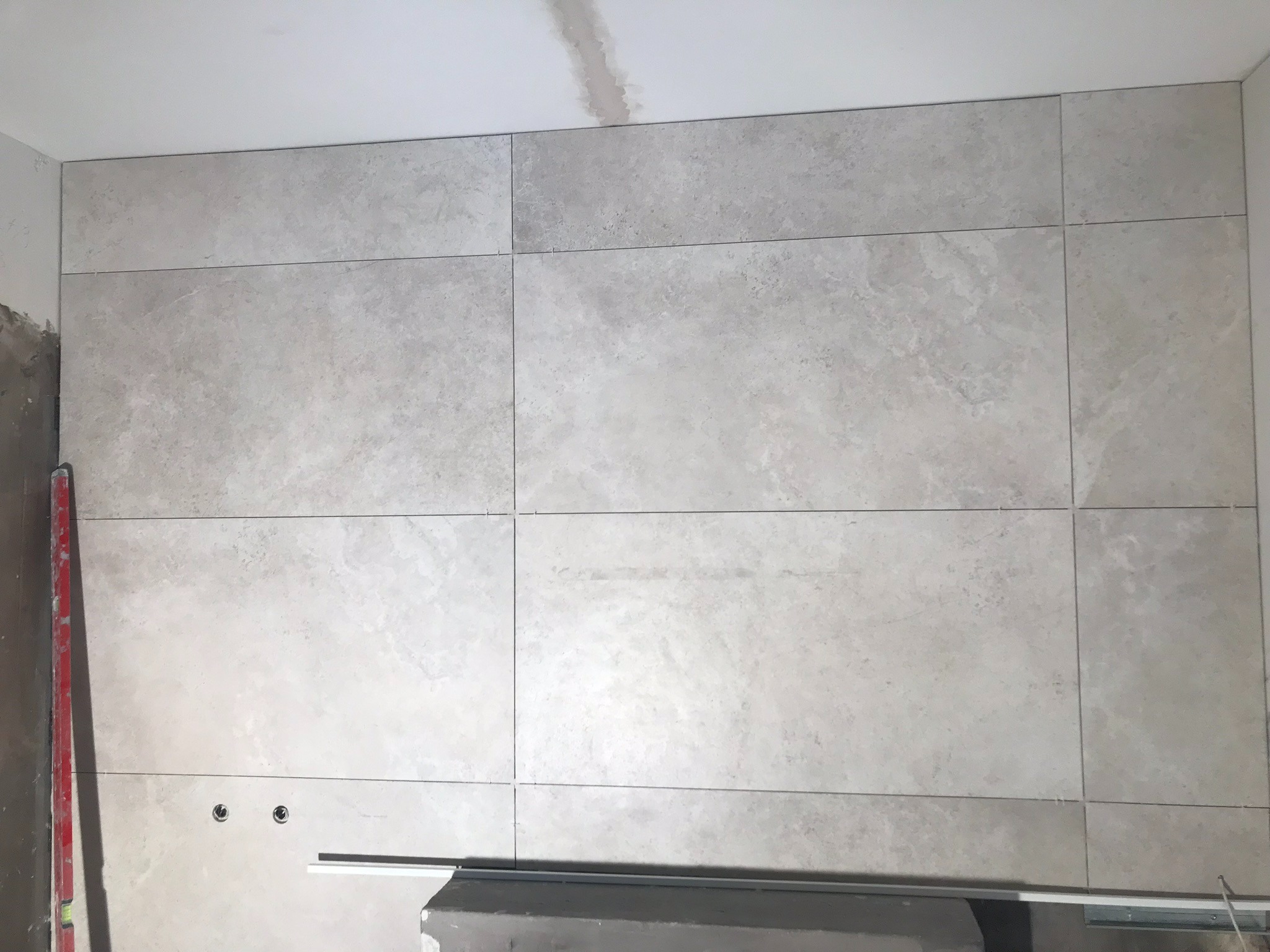 Ściana wyłożona jasnoszarymi płytkami ceramicznymi o matowej powierzchni, widoczne fugi, fragment poziomicy i element konstrukcyjny pod ścianą, dwa otwory na gniazdka elektryczne.