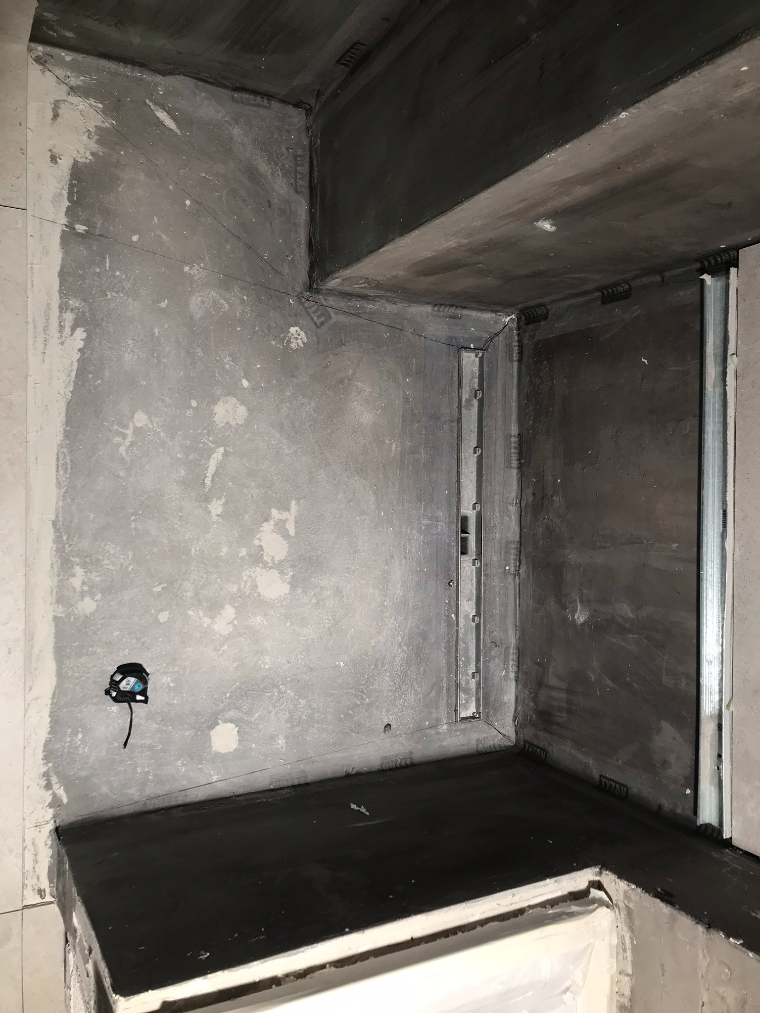 Surowa łazienka w trakcie remontu: ściany pokryte szarym tynkiem z widocznymi śladami po łatach, czarna hydroizolacja na podłodze i w narożnikach, zamontowany odpływ liniowy, widoczna puszka...