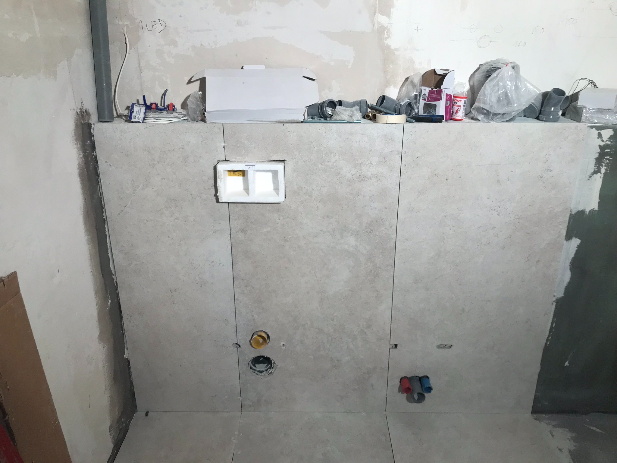 Ściana w trakcie układania płytek w łazience, z widocznymi otworami na instalacje wodno-kanalizacyjne oraz zabudową podtynkową toalety. Na górze ściany widoczne materiały budowlane i narzędzia.