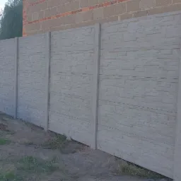 Długi, szary, betonowy płot z fakturą kamienia w Łąkcie, widoczny z bliska z niewielkim nasypem ziemi u podstawy.