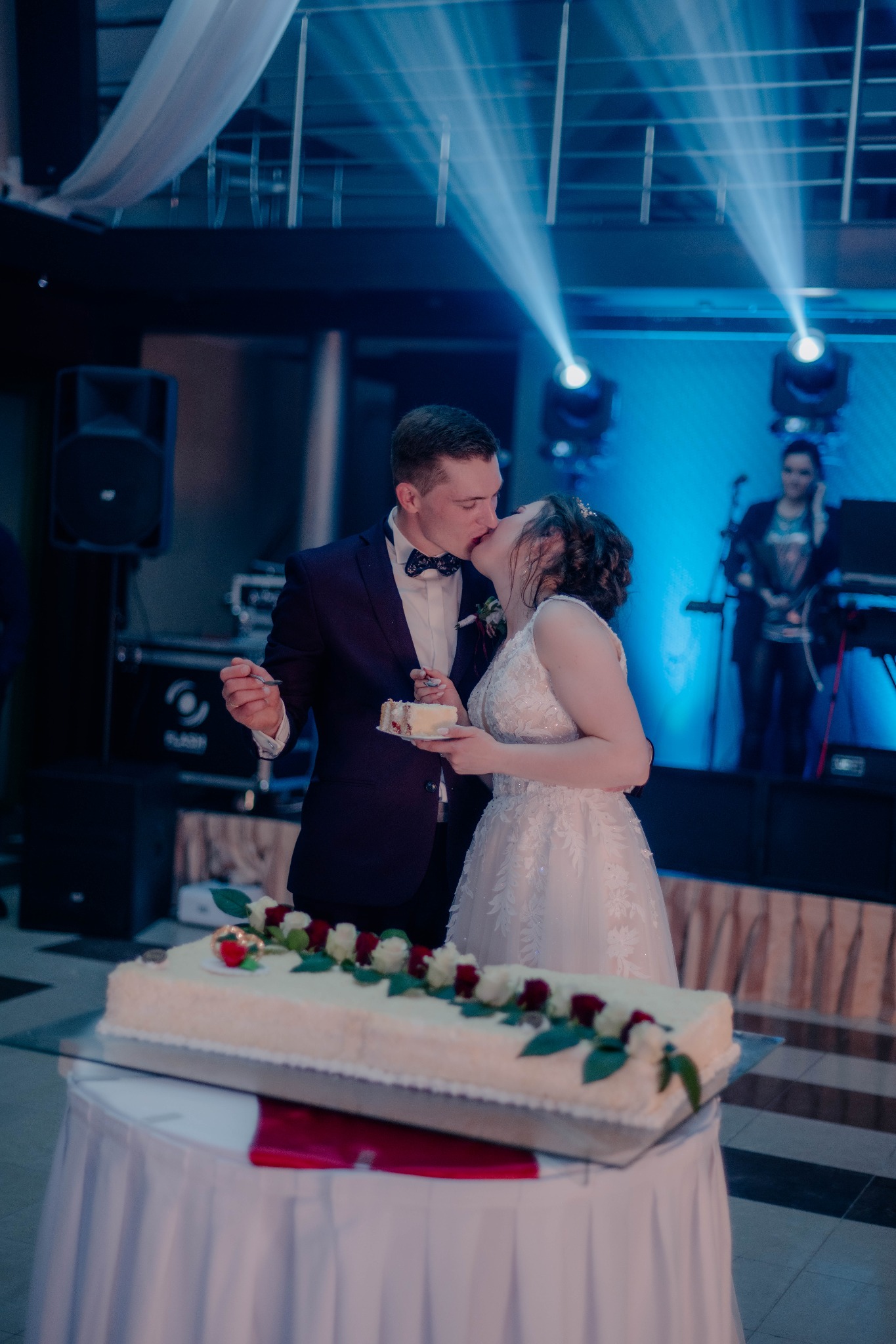 Nowożeńcy całują się, dzieląc tort weselny zdobiony białymi i czerwonymi różami, w tle scena z muzykami i reflektorami.