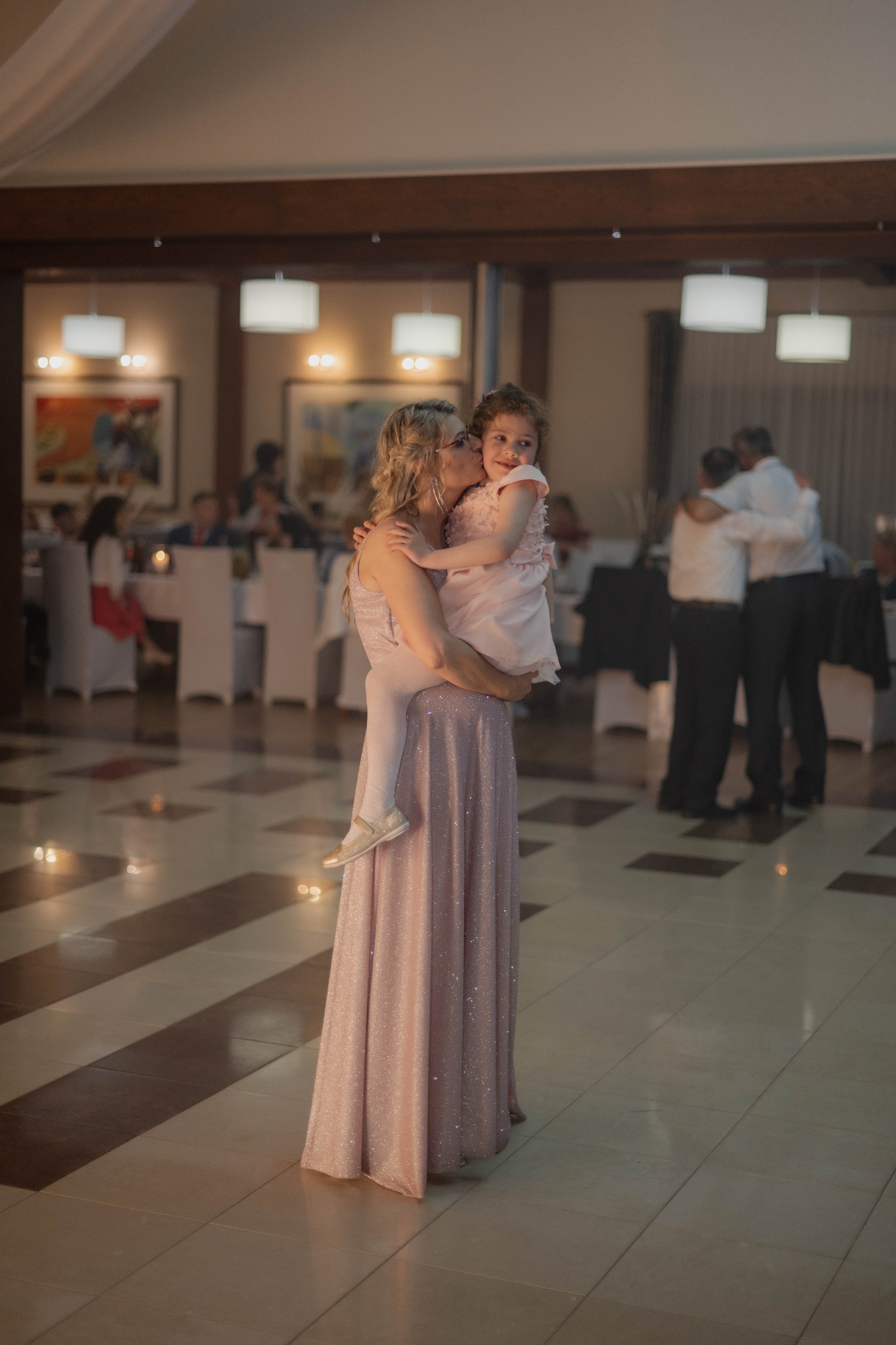 Kobieta w długiej, błyszczącej sukni trzyma na rękach dziewczynkę w różowej sukience, całując ją w policzek na sali balowej z innymi gośćmi w tle.