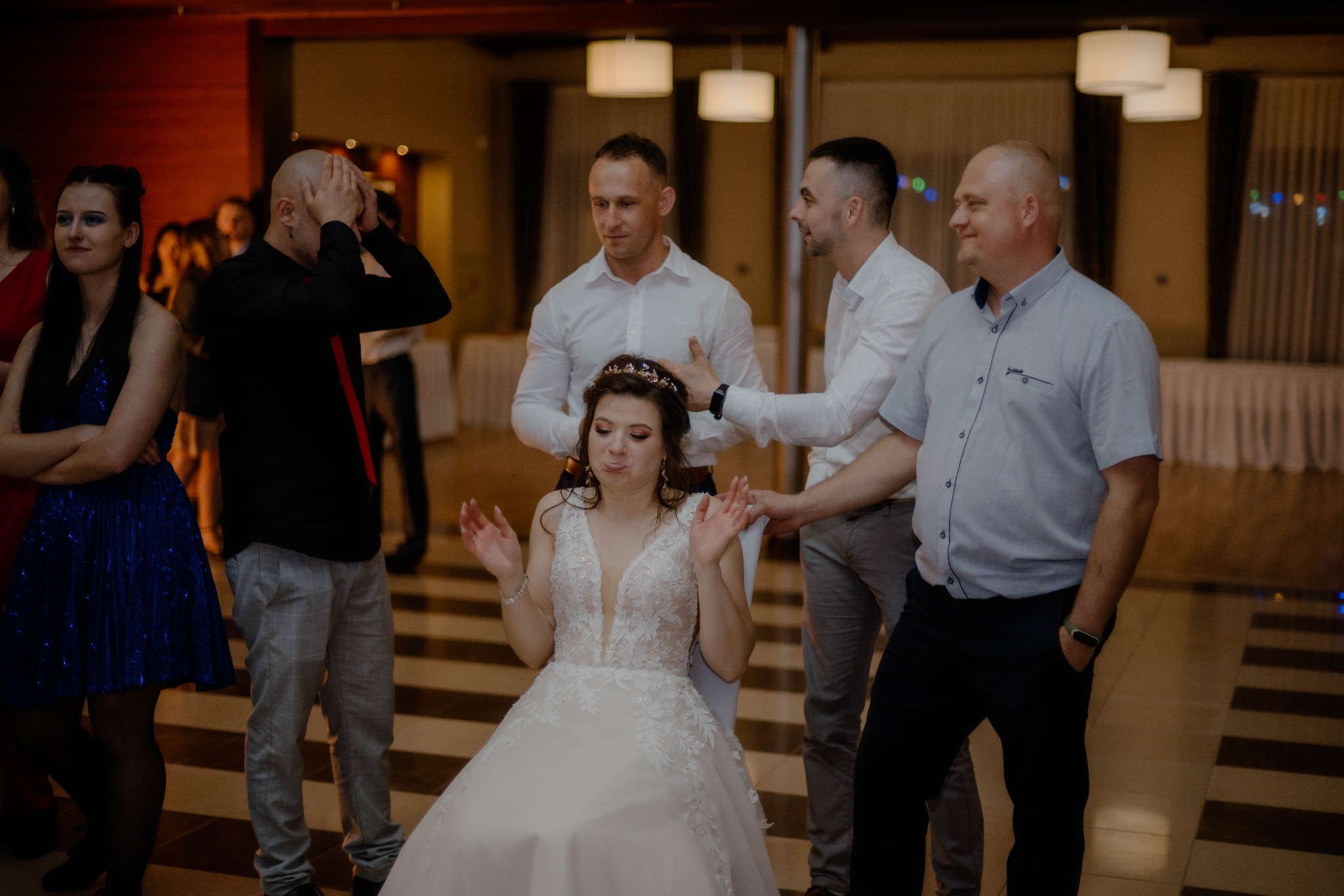 Panna młoda w białej sukni siedzi na krześle podczas oczepin, otoczona gośćmi weselnymi w swobodnych strojach, oświetlenie sali tworzy ciepłą atmosferę.