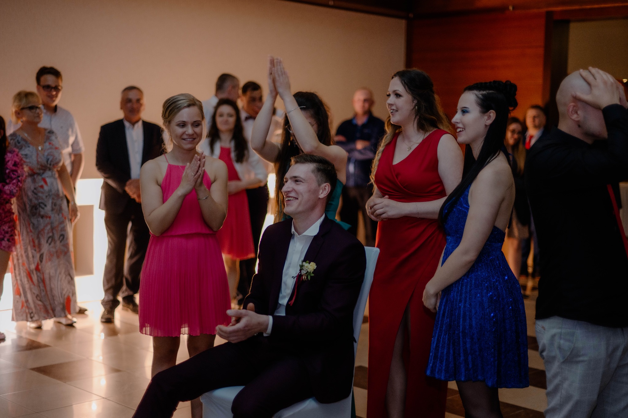 Uśmiechnięty mężczyzna w bordowym garniturze siedzi na krześle podczas uroczystości, otoczony przez gości w eleganckich strojach, w tle jasna ściana i parkiet.