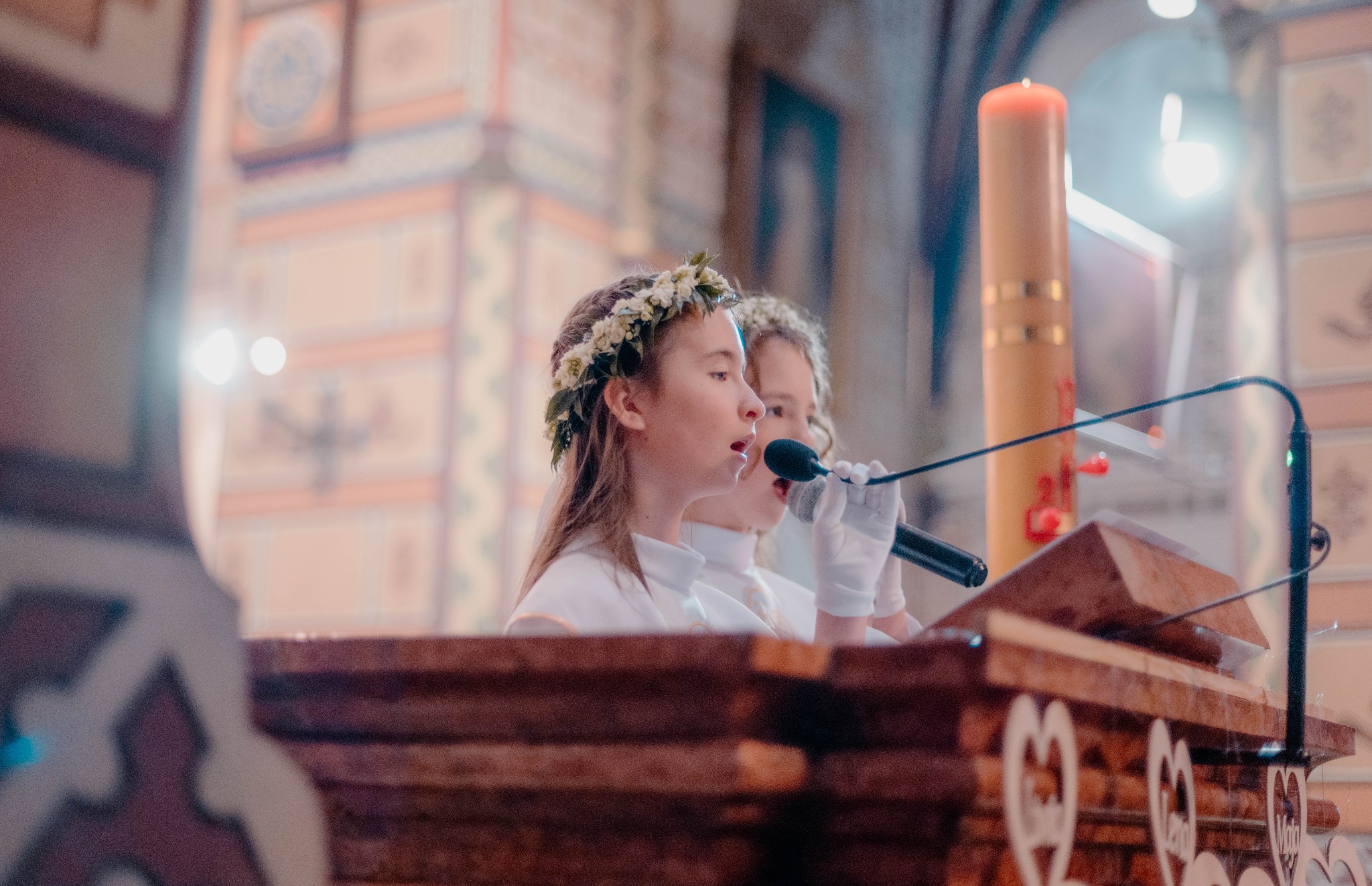 Dwie dziewczynki w białych strojach i wiankach na głowach śpiewają do mikrofonów podczas uroczystości w kościele, widoczna świeca paschalna i zdobiona ambona.