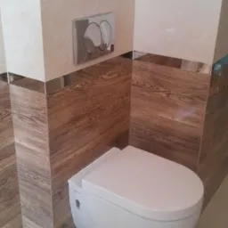 Zawieszona toaleta z zabudową w odcieniach beżu i brązu, z chromowanymi akcentami, widoczny przycisk spłukiwania i włącznik światła.