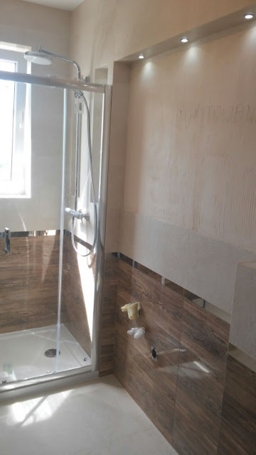 Wnętrze łazienki w trakcie remontu: kabina prysznicowa ze szklanymi drzwiami, ściany częściowo wyłożone płytkami imitującymi drewno, górna część ściany w stanie surowym, widoczne punkty świetlne...