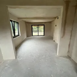 Wnętrze w stanie deweloperskim po nałożeniu gładzi gipsowej na ściany i wylewki cementowej na podłodze, widoczne okno z czarną ramą i przeszklone drzwi balkonowe.