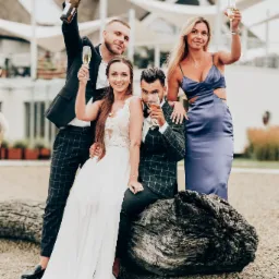 Uśmiechnięta para młoda siedzi na spr&oacute;chniałym pniu drzewa, wznosząc toast szampanem z dw&oacute;jką gości weselnych na tle nowoczesnej architektury.