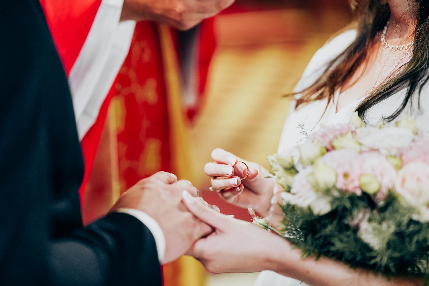 Wymiana złotych obrączek podczas ceremonii ślubnej, panna młoda trzyma bukiet z pastelowych kwiatów, w tle czerwono-złota szata liturgiczna.