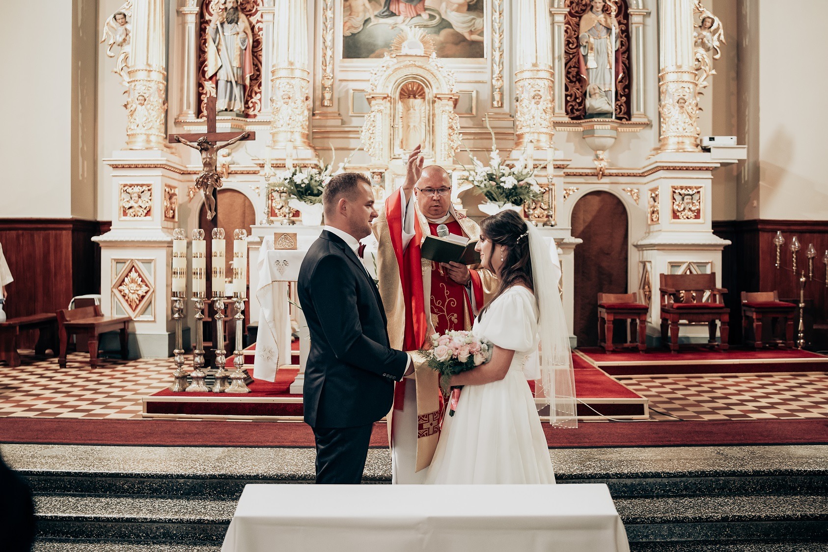 Para młoda podczas ceremonii ślubnej w bogato zdobionym kościele, ksiądz błogosławi z otwartą księgą.