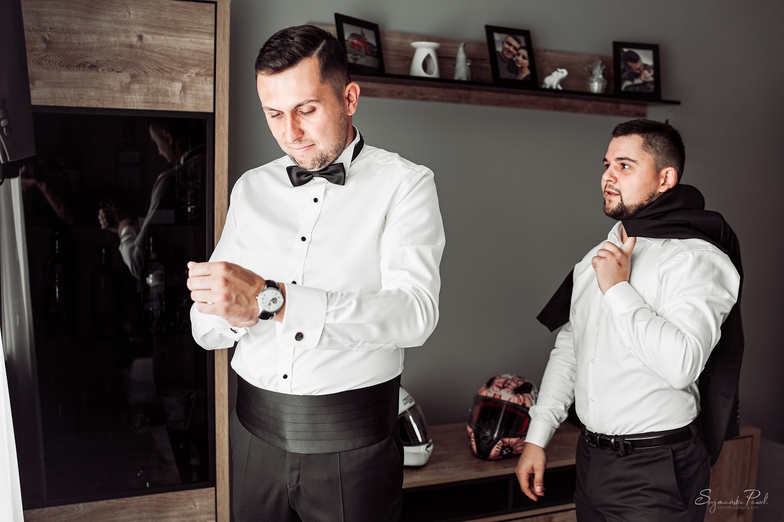 Pan młody w białej koszuli i czarnej muszce, poprawia zegarek na nadgarstku, obok stoi świadek z marynarką na ramieniu, w tle kask motocyklowy i zdjęcia w ramkach.