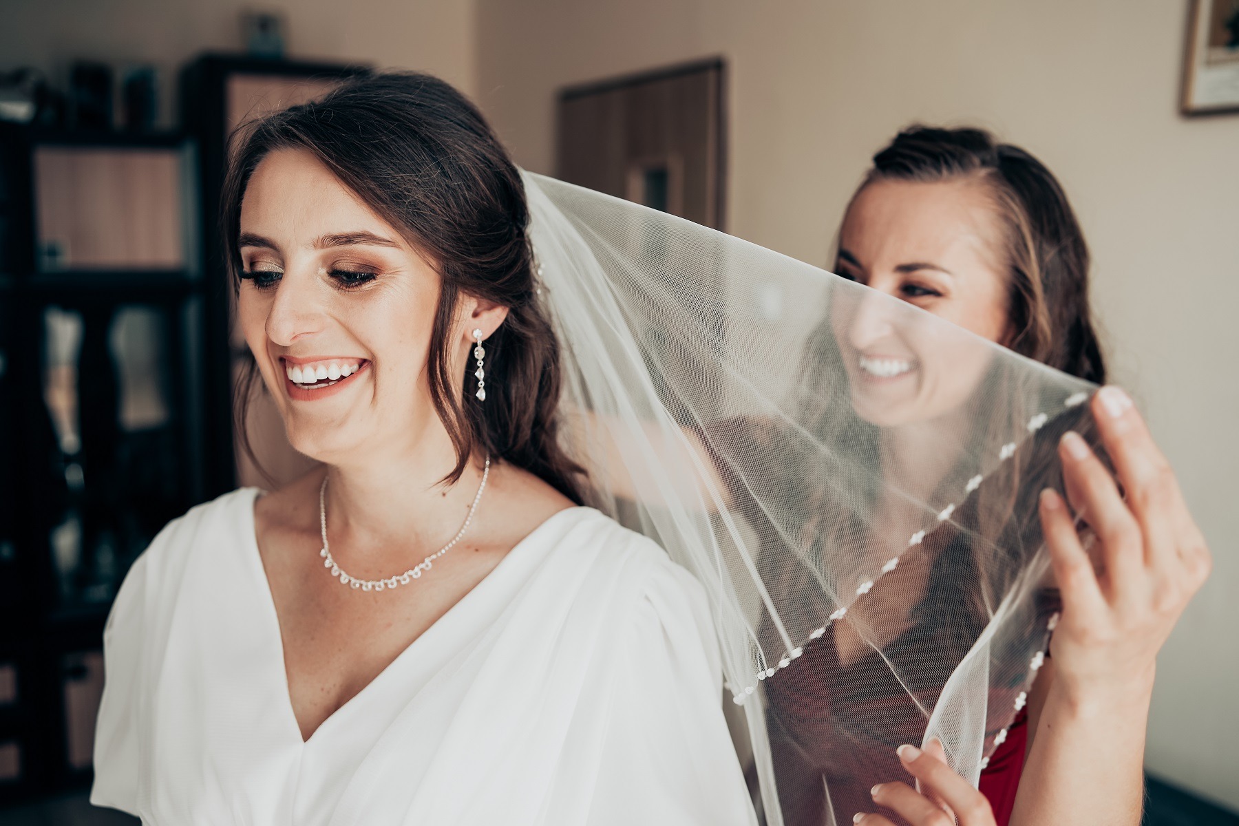 Uśmiechnięta panna młoda w białej sukni z dekoltem w serek, z delikatną biżuterią i makijażem, podczas zakładania welonu przez drugą kobietę.