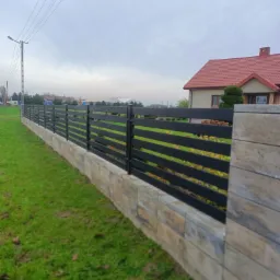 Nowoczesne ogrodzenie z poziomych paneli w kolorze antracytowym, osadzone na niskim murku z dekoracyjnych bloczków betonowych, z widokiem na dom z czerwoną dachówką w tle.