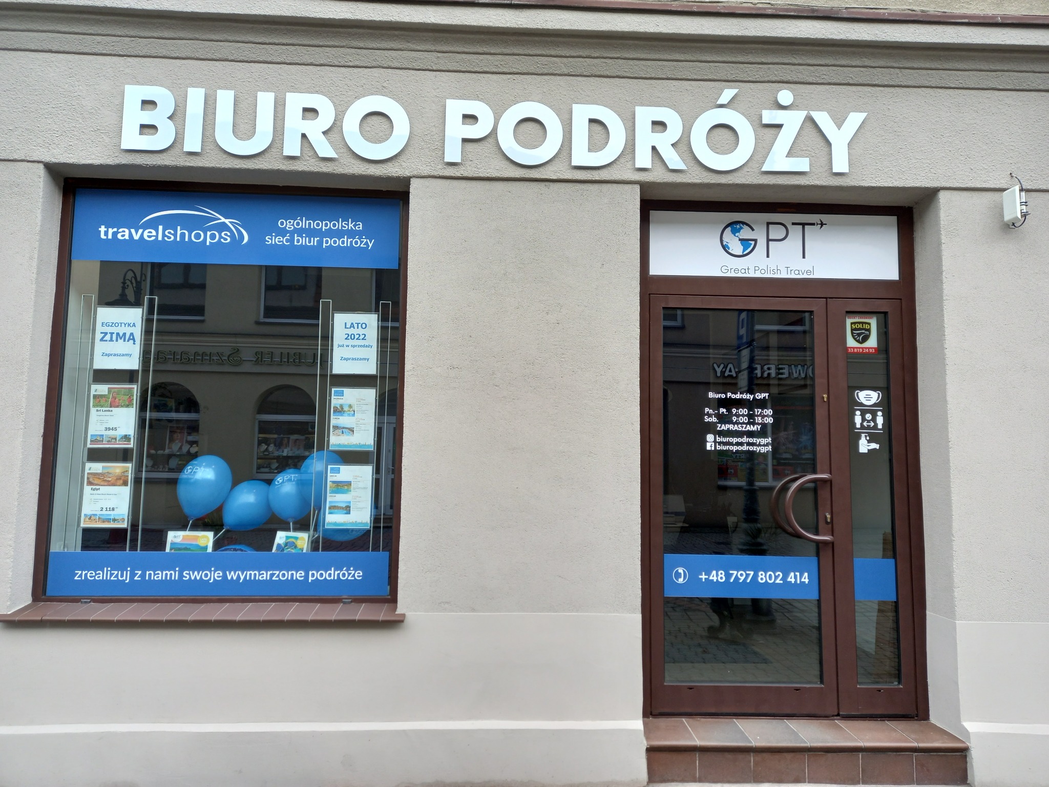 Fasada biura podróży z nazwą 'BIURO PODRÓŻY' nad witryną i drzwiami wejściowymi, oklejona plakatami ofert 'EGZOTYKA ZIMĄ' i 'LATO 2022', z niebieskimi balonami reklamowymi i numerem telefonu...