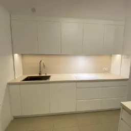 Nowoczesna, minimalistyczna kuchnia z białymi frontami, podświetleniem LED pod szafkami wiszącymi, zlewem z chromowaną baterią i płytą indukcyjną.