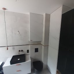 GutArt Radosław Gut - Nowoczesna łazienka z białą szafką pod umywalkę z czarną umywalką nablatową, wiszącą szafką i czarną toaletą, wykończona miedzianymi akcentami i oświetleniem, w trakcie montażu.