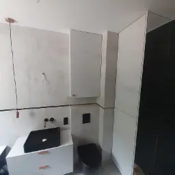 Nowoczesna łazienka z białą szafką pod umywalkę z czarną umywalką nablatową, wiszącą szafką i czarną toaletą, wykończona miedzianymi akcentami i oświetleniem, w trakcie montażu.