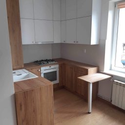 GutArt Radosław Gut - Jasna kuchnia z białymi szafkami górnymi i drewnopodobnymi szafkami dolnymi, z blatem i małym rozkładanym stolikiem przy oknie.